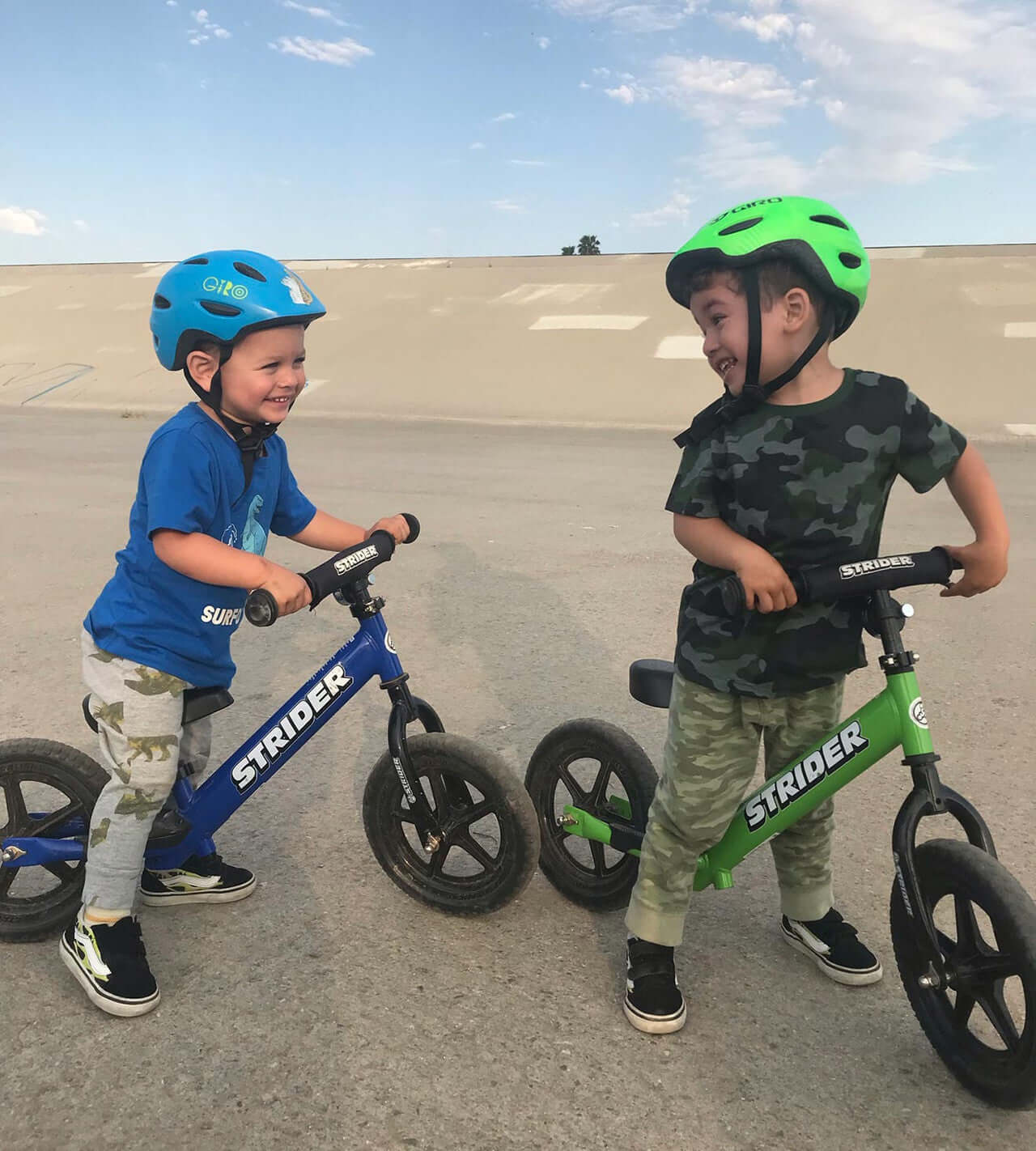 Two toddlers enjoying Strider 12 Sport Balance Bikes, wearing helmets and smiling, learning to ride outdoors.