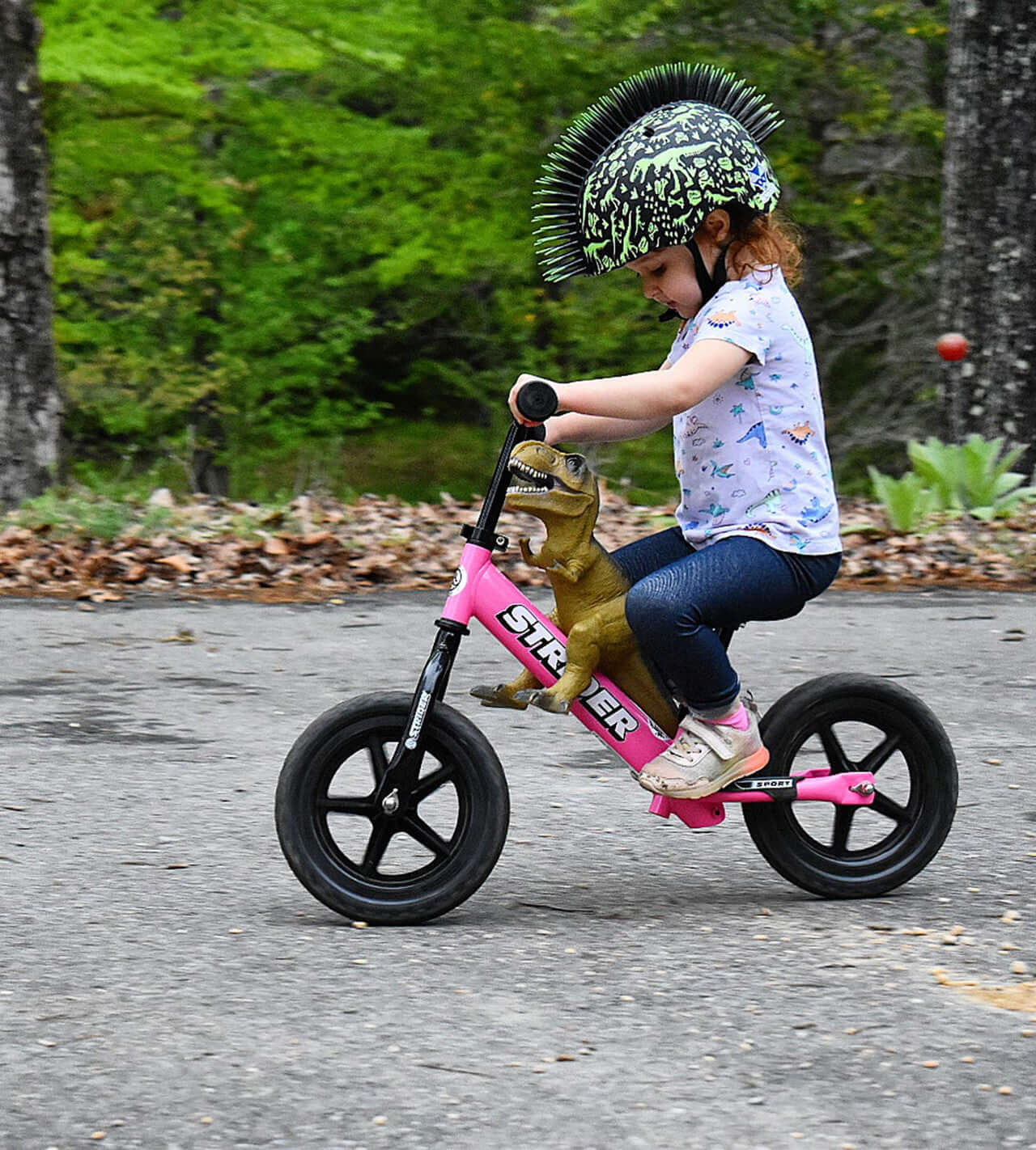 Child riding Strider 12 Sport Balance Bike with dinosaur plush toy and unique helmet in a wooded area.