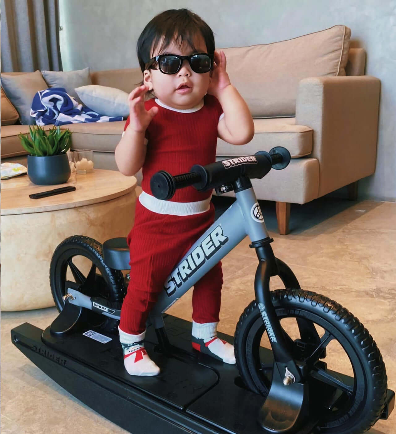 Baby on Strider Sport 2-in-1 Rocking Bike wearing sunglasses, learning to balance in a cozy living room.