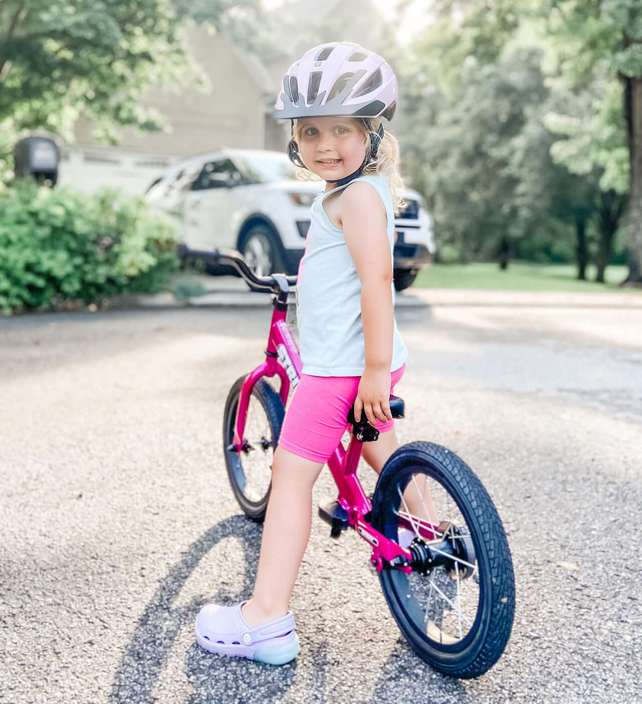 Smiling girl in a helmet riding a pink Strider 14x Convertible Bike on a sunny day, learning to balance and pedal.