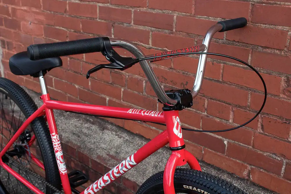 Close-up of the Eastern Growler BMX bike handlebars, featuring anodized grips and a sleek red frame against a brick wall.