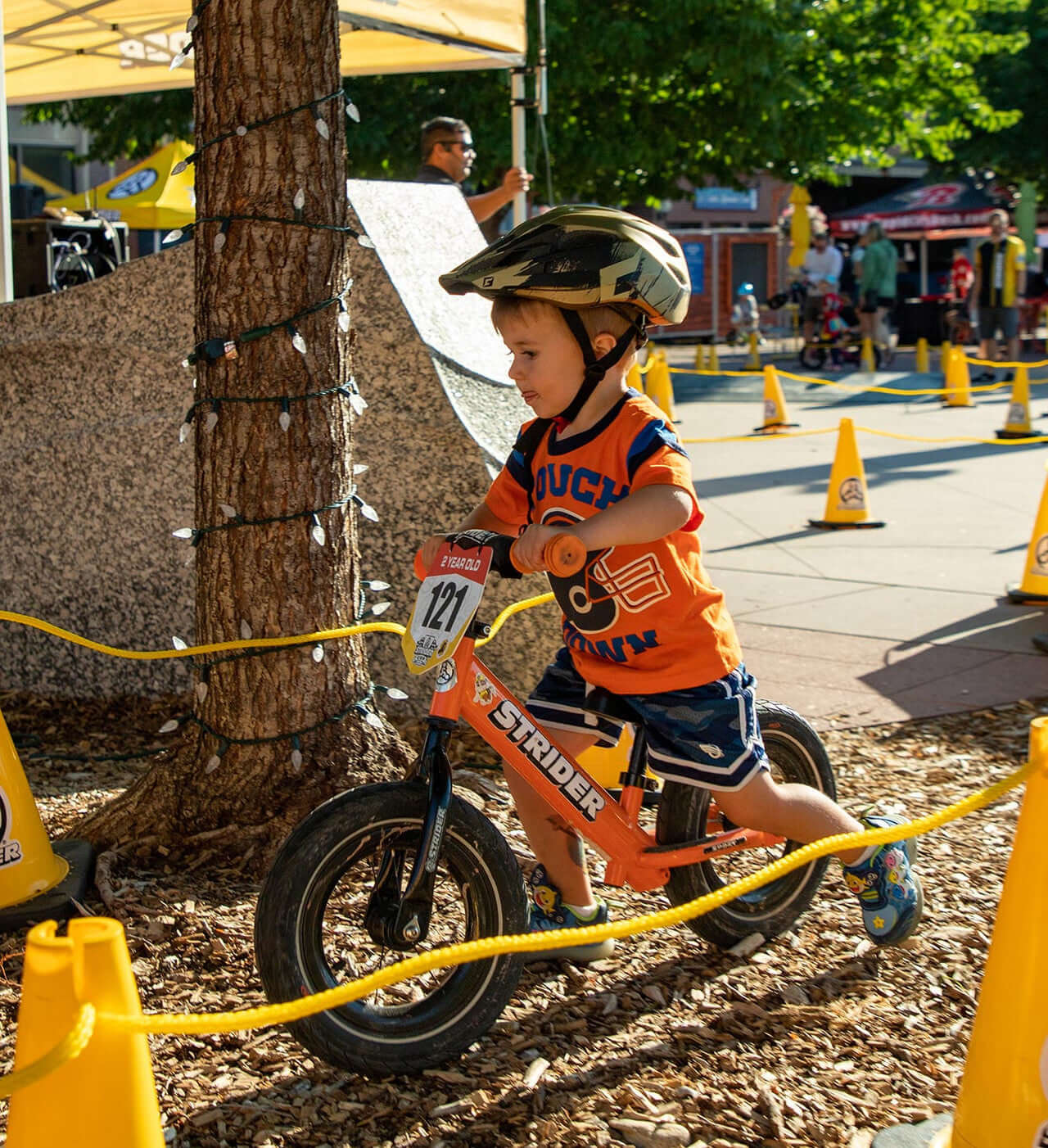 Young child racing on a Strider Balance Bike, wearing a helmet and orange shirt, in a vibrant outdoor setting.