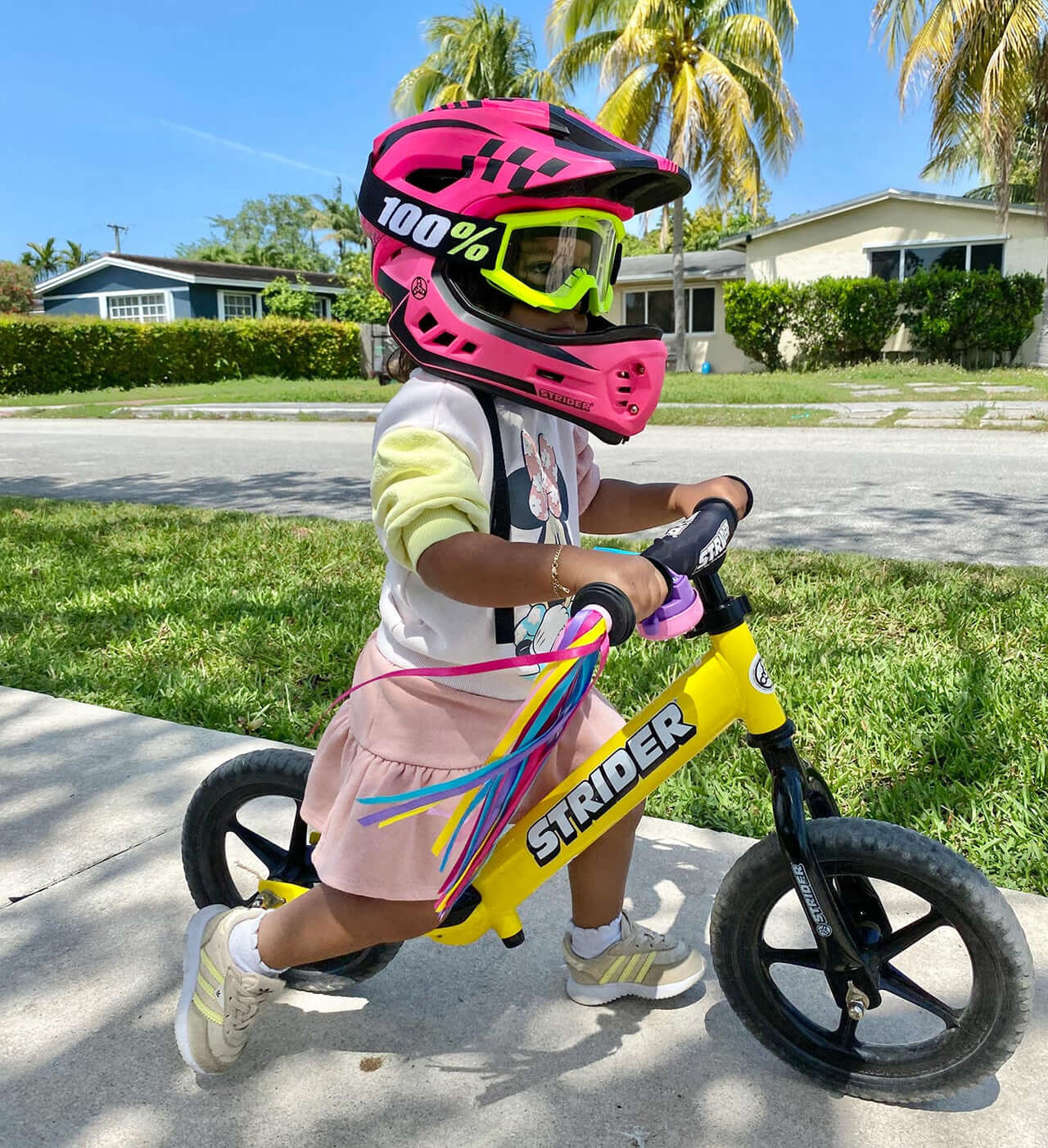 Child wearing a pink Strider 2-in-1 full face helmet while riding a yellow Strider balance bike outside.