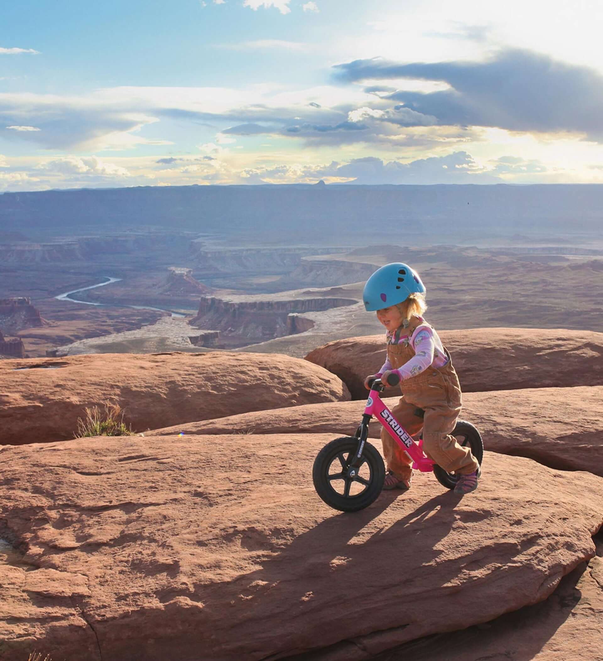 Child riding a Strider balance bike on rocky terrain with a scenic landscape in the background.